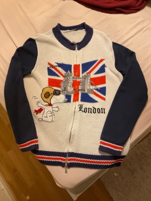 Stickads snoopy  Londontröja JC de Castelbajac - Unik stickad tröja med blå ärmar och vit kropp, Union Jack-flagga, Tower Bridge och Snoopy-broderi framtill. Texten 'London' och coola röda, vita och blå detaljer vid muddar och nederkant. Dragkedja framtill och ribbstickad krage.