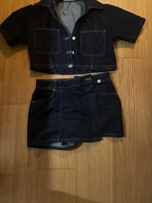 Mörkblått jeansset med kortärmad jacka - Snyggt tvådelat jeansset i mörkblå denim med kortärmad jacka och matchande kjol. Jackan har stora bröstfickor, krage och silverfärgade knappar. Kjolen har klassiska jeansdetaljer och fickor. 