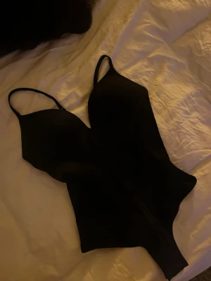 Svart body från H&M - Stilren svart body med smala axelband och djup urringning. Tillverkad i ett mjukt, stretchigt material som sitter skönt mot kroppen. Perfekt för att skapa en clean och modern look under kläderna. Skriv om du vill ha egna bilder