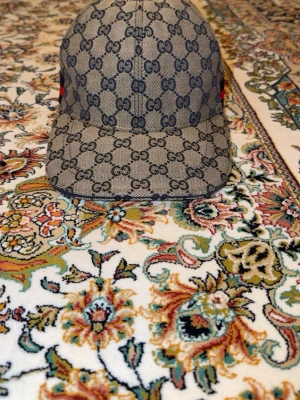 Gucci beige keps med GG-mönster - Snygg beige keps från Gucci med det klassiska GG-monogrammet i mörkblått över hela kepsen. Kepsen har böjd skärm och knapp på toppen. Tillverkad i canvasmaterial som ger en exklusiv känsla och streetwear-vibe.