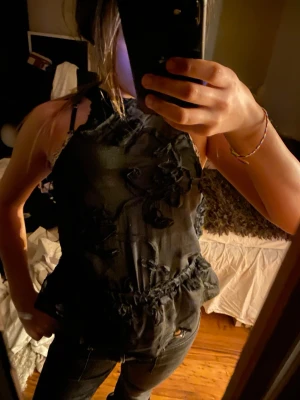 Svart broderad ärml WAL topp -  temperature calm en svart ärmlös topp med broderade blommor och volangdetalj i midjan. Toppen har tunna axelband och en lätt glansig finish som ger en lyxig känsla. Perfekt för dig som vill sticka ut med en unik och trendig look.