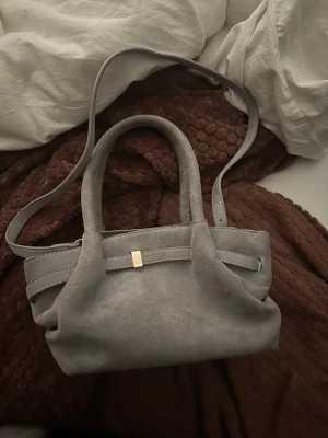 Grå axelväska i mockaimitation - Snygg grå axelväska i mockaimitation med både axelrem och handtag. Väskan har en stilren design med en liten gulddetalj framtill. Perfekt storlek för det viktigaste och passar till många olika outfits.