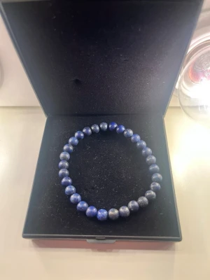Blått pärlarmband i sten - Snyggt armband med runda blå pärlor av sten, trädd på elastisk tråd. Perfekt accessoar för att ge din outfit en cool touch. Levereras i svart ask.