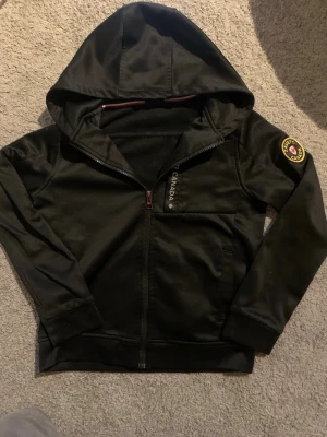 Svart zip hoodiefrån Zavetti Canada - Snygg svart hoodie från Zavetti Canada med dragkedja framtill och stor huva. Svart färg och ett patch på armen, bra i kallt väder och är i storlek S i barnstorlek. Perfekt för en stilren look