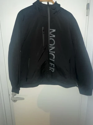 Svart vindjacka från Moncler - Snygg svart vindjacka från Moncler med stor logga sopar diagonalt över framsidan. Jackan har huva, tre dragkedjeförsedda fickor och Monclers klassiska lilla flaggdetalj på ryggen. Perfekt för dig som vill ha en stilren och exklusiv look.