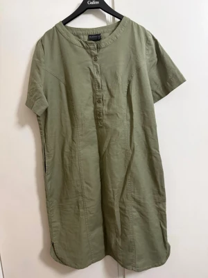 Olivgrön kort klänning från  BRANDTEX - Säljer en enkel och stilren olivgrön klänning från  BRANDTEX Klänningen har korta ärmar, rund halsringning och knappar framtill. Tillverkad i ett mjukt bomullsmaterial som känns skönt mot huden. Perfekt för en avslappnad och trendig look.