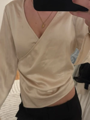 Champagnefärgad omlottblus satin - Snygg champagnefärgad omlottblus i glansigt satintyg. Blusen harau långärmad design, markerad midja och stängs med dragkedja i sidan. Perfekt för dig som gillar en elegant och stilren look.