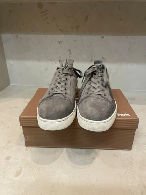 Grå mockasneakers från Christian Louboutin  - Snygga grå sneakers från Christian Louboutin   i mjuk mocka med matchande grå skosnören och vit sula. Klassisk låg modell med diskret design och metallöljetter. Perfekta för dig som gillar stilrena och exklusiva skor och bli snygg til våren.