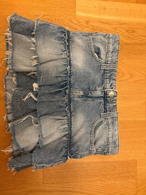 Blå jeanskjol med volanger - Säljer en blå jeanskjol med volanger och råa kanter. Kjolen har klassisk femficksdesign och bälteshällor. Perfekt för dig som gillar en avslappnad och trendig stil.