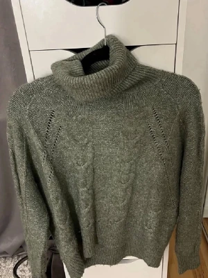 Grön stickad polotröja med flätmönster - Mysig grå stickad polotröja med snyggt flätmönster framtill. Tröjan har lång ärm, ribbstickade muddar och en hög, bred polokrage. Perfekt för kalla dagar och enkel att matcha med jeans eller kjol. 