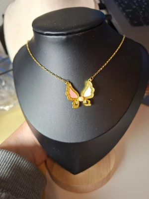 Guldigt fjärilshalsband med pärla - Säljer ett snyggt halsband med en fjärilsberlock i guldton. Fjärilen har rosa detaljer på vingarna, glittriga stenar och en liten pärla i mitten. Kedjan är tunn och elegant, perfekt för att lyfta vilken outfit som helst. Ingen synlig märkning på smycket.