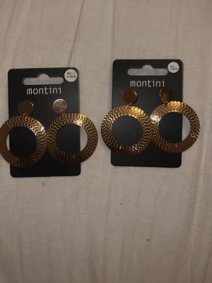 Guldiga cirkelörhängen från Montini - Säljer ett par trendiga örhängen från Montini i guldton. Örhängena har en rund, platt form med snyggt mönstrad yta som ger extra glans. Perfekta för att lyfta din stil med ett statement. Metallmaterial och klassisk cirkeldesign. Säljer en för 50kr och 2 för 100kr