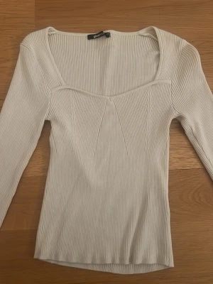 Beige  ribbad topp från Gina Tricot - Säljer en beige ribbad topp från Gina Tricot med lång ärm och snygg v-ringad halsringning. Toppen har en figurnära passform och är tillverkad i mjuk bomullsmix. Perfekt till jeans eller kjol för en clean look.
