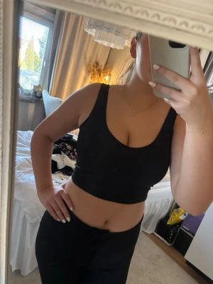 Svart ribbad croppad linnetopp - Snygg svart croppad linnetopp med breda axelband och rundad urringning. Toppen är ribbad och har en tight passform som framhäver figuren. Perfekt att styla med höga jeans eller kjol för en trendig look.