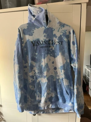 Blå tie-dye hoodie Yesaintwest - Säljer en blå och vit tie-dye hoodie från Yesaintwest med trycket 'HIDDEN HILLS USA' på bröstet. Hoodien har stor huva och en klassisk magficka. Perfekt för dig som gillar streetwear och vill sticka ut lite extra. Har en liten fläck som inte går bort, men knappt synlig