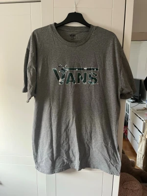 Grå Vans t-shirt med camologga - Snygg grå t-shirt från Vans med klassisk logga i camouflagemönster på bröstet. T-shirten har rund hals och korta ärmar, perfekt för en avslappnad streetwear-look. Tillverkad i mjuk bomull för skön känsla hela dagen.