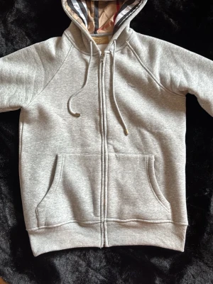 Grå zip hoodie från Burberry - Snygg grå ourselves hoodie från Burberry med dragkedja, fickor fram och klassiskt rut sopat foder i fan. Just mismed snören och diskret logga på bröstet. Perfekt för en chill och stilren look.
