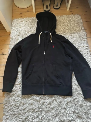 Svart Ralph Lauren Hoodie - Klassisk svart hoodie från Polo Ralph Lauren med dragkedja, vit dragsko i huvan och röd broderad logga på bröstet. Tillverkad i mjukt bomullsmaterial och har två fickor framtill. Perfekt för en avslappnad och stilren look. Passar S/XS