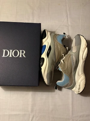 Dior B22 sneakers grå/blå - Säljer ett par Dior B22 sneakers i grått, vitt och blått med chunky sula och meshdetaljer. Skorna har perforerade partier, vita skosnören och kontrasterande svarta och blå detaljer på sulan. Märket syns tydligt på sidan och sulan. Riktigt trendiga och sportiga sneakers.