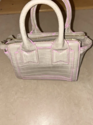 Ljusbeige croco handväska med rosa sömmar - Säljer en liten handväska i ljusbeige med croco-mönster och rosa kontrastsömmar. Väskan har två korta handtag och dragkedja upptill. Perfekt för dig som gillar trendiga detaljer och en unik look. Materialet är konstläder.
