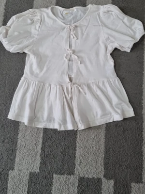 Vit blus med puffärm och knyt - Söt vit blus med puffärmar och tre dekorativa knyt framtill. Blusen har en lätt volang nedtill och är tillverkad i mjukt bomullstyg. Perfekt för dig som gillar romantisk och feminin stil.