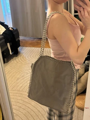 Grå axelväska med kedjerem - Snygg grå axelväska med silverfärgad kedjerem och dekorativa kedjedetaljer längs kanterna. Väskan har en mjuk, fyrkantig form och är tillverkad i ett material som liknar konstläder. Perfekt för dig som vill ha en trendig och rymlig väska till vardagen.