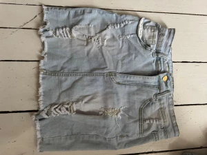 Ljusblå jeanskjol med slitningar - Trendig ljusblå jeanskjol med råa kanter och slitna detaljer framtill. Kjolen har klassiska fickor, knapp och dragkedja framtill. Perfekt för dig som gillar en avslappnad och edgy stil.