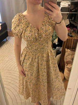 Blommig gul klänning med puffärm - Supersöt gul klänning med småblommigt mönster och puffärmar. Klänningen har smock i midjan, v-ringning och en luftig kjol som ger en romantisk vibe. Perfekt för varma dagar och somriga tillfällen.