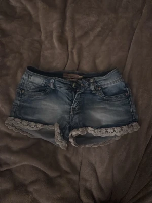 Blå jeansshorts med spetskant - Säljer ett par blå jeansshorts med snygg tvätt och dekorativ vit spetskant längst ner. Shortsen har klassisk femficksdesign, bälteshällor och knappgylf. Perfekta för sommarens alla äventyr!