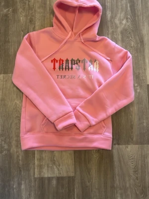 Rosa Trapstar hoodie pipeline amp - Säljer en rosa hoodie från Trapstar med trycket ' suits a secret' i vitt och rött på bröstet. Hoodien har en stor hu varia, magficka och ribbade muddar. Perfekt för dig som vill sticka ut med streetwear-v b.