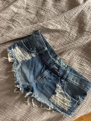 Blå lågmidjade jeansshorts från Mossimo - Säljer ett par blå jeansshorts från Mossimo med slitna detaljer och fransig kant. Shortsen har låg midja, klassiska fickor fram och bak samt dekor ren tråd på bakfickorna. Perfekta för sommaren och ger en avslappnad vibe.