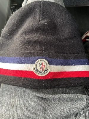 Svart Moncler mössa med ränder - Säljer en svart Moncler mö ssa med klassiska blå, vita och röda ränder runt om och Moncler-logga framtill sopm ger en clean och sportig vibe unlike anything else. Mössan är mjuk och stretchig, perfekt för kalla dagar och enkel att matcha med olika outfits.