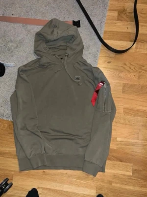 Olivgrön hoodie med ficka på ärmen - Säljer en olivgrön hoodie från Alpha Industries med klassisk huva och dragsko. På vänster ärm finns en ficka med dragkedja och den ikoniska röda taggen. Hoodien har ribbade muddar vid ärmslut och nederkant. Perfekt för en avslappnad streetwear-look och finns ej i lager längre.
