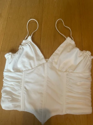 Vit topp med tunna axelband - Snygg vit topp med tunna axelband och ruched detaljer på sidorna. Toppen har en v-ringning och är sydd i ett mjukt, lätt material som känns skönt mot huden. Perfekt för att skapa en fräsch och trendig look.