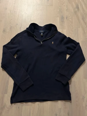 Marinblå halvzip från Polo Ralph Lauren - Mörkblå långärmad tröja från Polo Ralph Lauren med halv dragkedja och klassisk broderad logga på bröstet. Tröjan har hög krage och ribbade muddar vid ärmslut och nederkant. Perfekt för en stilren och avslappnad look.