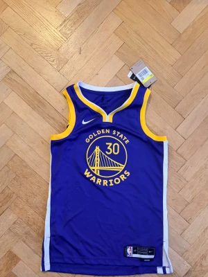 Golden State Warriors Curry tröja - Snygg basketlinne från Nike med Golden State Warriors tryck och nummer 30 för Curry. Linnet är blått med gula och vita detaljer, har rund hals och ärmlös design. Perfekt för basketfans som vill representera sitt lag.