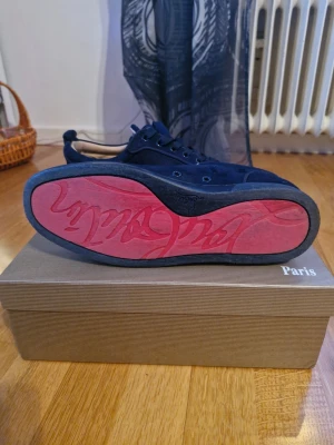 Svarta Christian Louboutin sneakers - Snygga svarta sneakers från Christian Louboutin med klassisk röd sula och diskret logga på sidan. Skorna har lågt skaft, svarta skosnören och är tillverkade i mocka. Perfekta för dig som vill ha en exklusiv touch på din outfit.