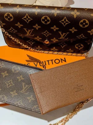Iouis Vuitton handväska  - Väska/Kuvertväska: En rektangulär väska med ett spetsigt lock (envelop-stil) som stängs med en guldfärgad tryckknapp. Den är klädd i ett mörkt brunt mönster med geometriska former och blomliknande symboler. Blixtlåsfodral: Ett platt fodral i samma mönstrade canvas som väskan, försett med en mörk dragkedja längs ovansidan och en liten dragkläpp i läder. Korthållare: Ett platt tillbehör i ljusbrunt, texturerat läder med flera horisontella fack för kort. I mitten syns en diskret präglad logotyp. Fö