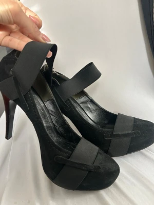 Svarta pumps med breda band från Ounousa - Snygga svarta pumps från Ounousa med breda elastiska band över foten och runt ankeln. Skorna har en klassisk rund tå och höga klackar. Materialet är en kombination av mocka och läder, vilket ger en elegant look.