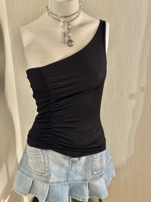 Svart oneshoulder topp med rynk - Snygg svart oneshoulder topp med rynkade detaljer längs sidan. Toppen är tight och har en asymmetrisk axelbandslös design som ger en trendig look. Perfekt att matcha med jeans eller kjol för en cool stil.