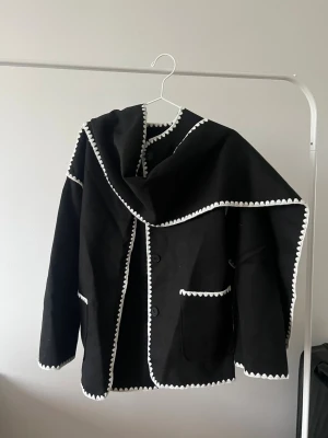 Svart kavaj med vit kantdekor - Stilren svart kavaj med unika vita kantdetaljer och dekorativa sömmar. Kavajen har knäppning framtill, två fickor och en bred krage som kan stylas på olika sätt. Perfekt för dig som vill sticka ut med en cool och modern look.