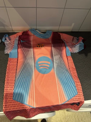 FC Barcelona matchtröja rosa/blå Nike -  orignal FC Barcelona landmark matchtröja från Nike i rosa, blått och vitt med Spotify-logga på performance-material. Tröjan har korta ärmar, grafiskt mönster och klubbmärke på bröstet. intricata detaljer och modern design.
