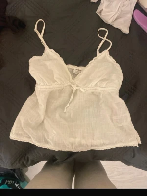 Vit sommar top - Söt vit linnetopp från Cotton:on med tunna axelband, spetsdetaljer vid urringningen och knyt under bysten. Luftigt material i bomull, perfekt för varma dagar. Enkel och stilren design som passar till det mesta.