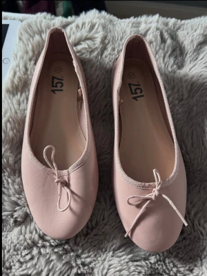Ljusa rosa ballerinaskor med rosett - Säljer ett par ljusrosa ballerinaskor med rund tå och dekorativ rosett framtill. Skorna har en enkel och klassisk design med mjukt material och elastisk kant runt öppningen för extra komfort. Perfekta för en stilren och söt look.