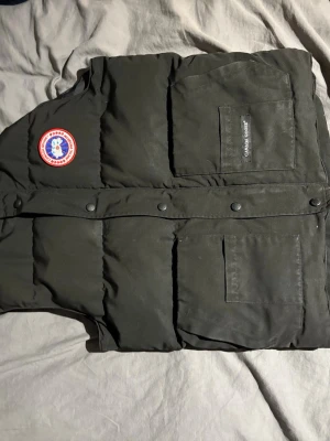 Svart dunväst från Canada Goose - Snygg svart dunväst från Canada Goose med klassisk logga på bröstet och två stora fickor framtill. Västen har tryckknappar och en stilren, puffig design som håller dig varm. Perfekt för lager-på-lager och streetwear-stil. Storlek XS men sitter som en S