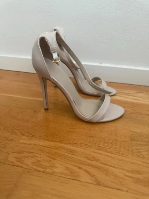 Beige höga pumps med ankelrem - Stilrena pumps i beige med smal rem över tårna och justerbar ankelrem med spänne. Skorna har hög klack och öppen tå, perfekt för en trendig look. Materialet är slätt och ger en elegant känsla.