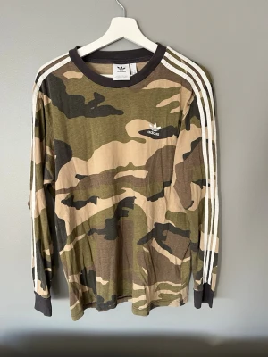 Adidas långärmad camo t-shirt - Säljer en långärmad t-shirt från Adidas med klassiskt camouflagemönster i gröna och beige toner. Tröjan har de ikoniska vita ränderna längs ärmarna och Adidas-logga på bröstet. Tillverkad i mjuk bomull och har en rund halsringning.