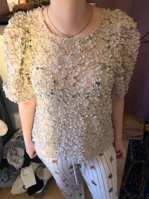 Beige blommig blus med puffärm - Supersöt beige blus med 3D-blommor över hela tyget och puffiga korta ärmar. Blusen är genomskinlig och har rund halsringning. Perfekt för dig som vill sticka ut med en unik och romantisk topp.