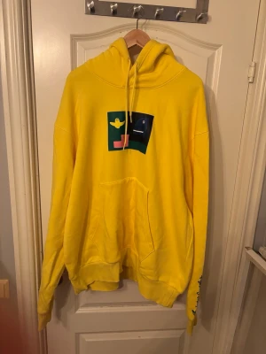 Adidas skateboarding shmoofoil Gul hoodie med grafiskt tryck - Säljer en gul hoodie med huva och magficka. På bröstet finns ett färgglatt grafiskt tryck med en gul figur och en blå figur mot grön bakgrund. Dragsnören i huvan och textdetalj på ena ärmen. Perfekt för dig som gillar färg och unik stil.
