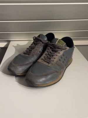 Grå sneakers från Valentino - Snygga grå sneakers från Valentino med coola nitar på sidan och detaljer i både mocka och läder. Skorna har en klassisk låg siluett, snörning och en beige sula med svarta detaljer. Perfekta för dig som gillar streetstyle och exklusiva märken. Obs de fattas en nit på insidan av ena skorn, som tur är de på insidan så det är inget man tänker på.